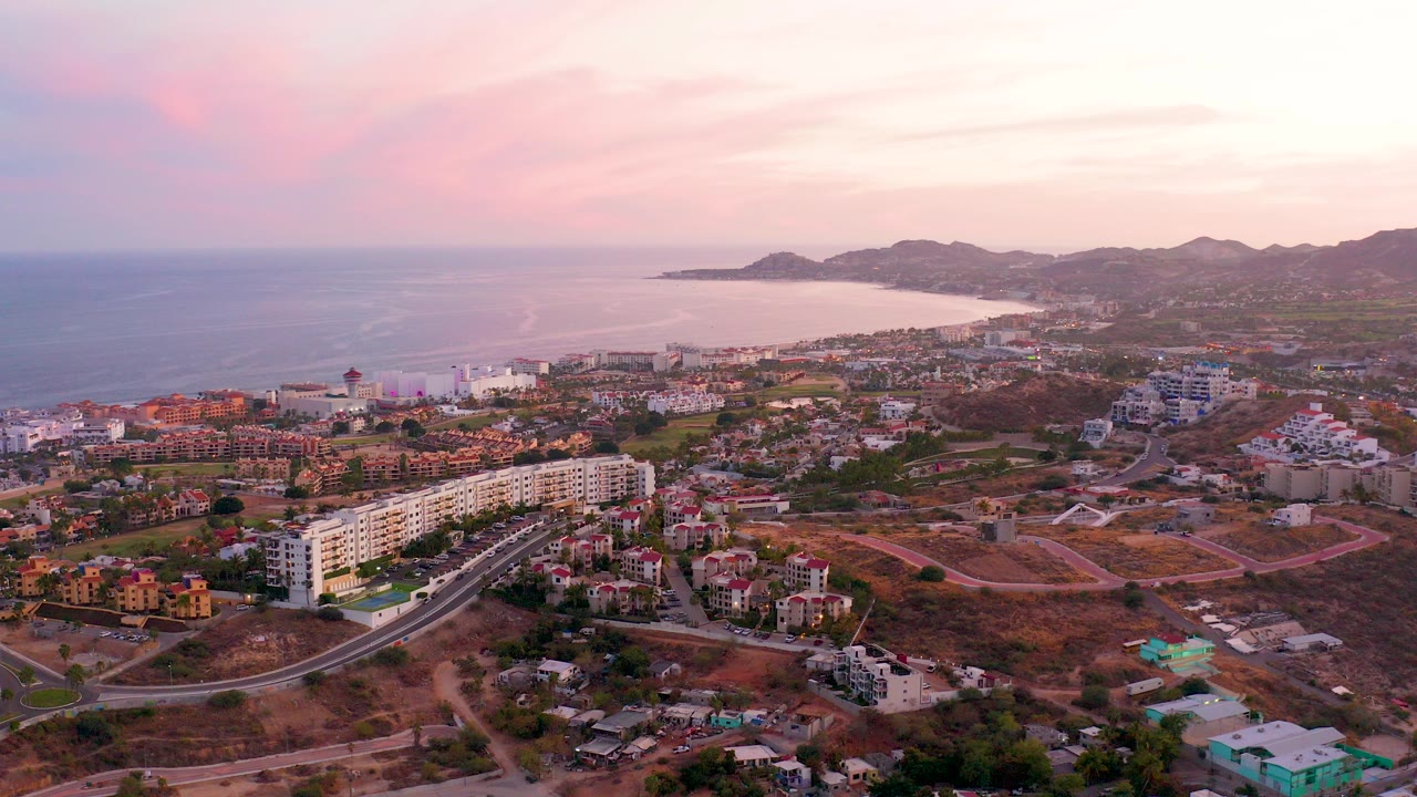 Wide aerial view over San Jose del Cabo, Mexico, with low-rise buildings, a curving road, dry lots, ocean water, distant hills, pink evening clouds, and no visible people.