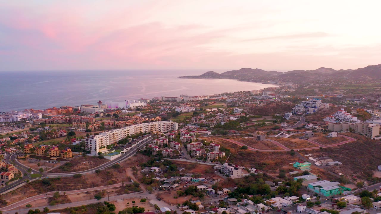 Aerial view over San Jose Del Cabo with residential buildings, curving roads, ocean to the left, distant hills, and a pink dusk sky, with no visible people.