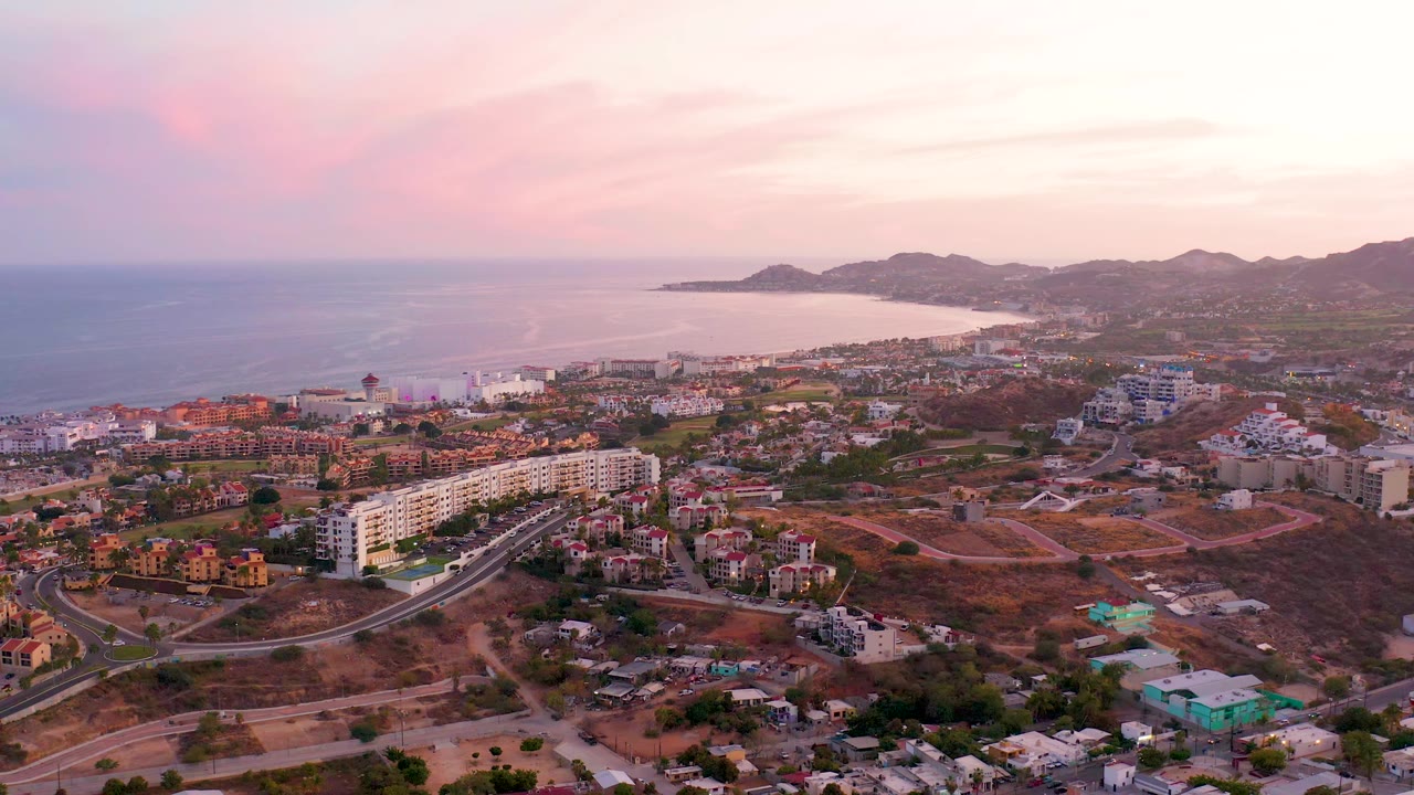 San Jose Del Cabo appears from above with apartment blocks and low homes in the foreground, the ocean across the left background, and hills under pink evening light, with no visible people.