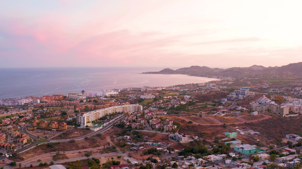 Wide opening aerial over San Jose del Cabo, Mexico, showing hillside roads, residential buildings, the ocean, distant mountains, and a pink sky with no visible people.