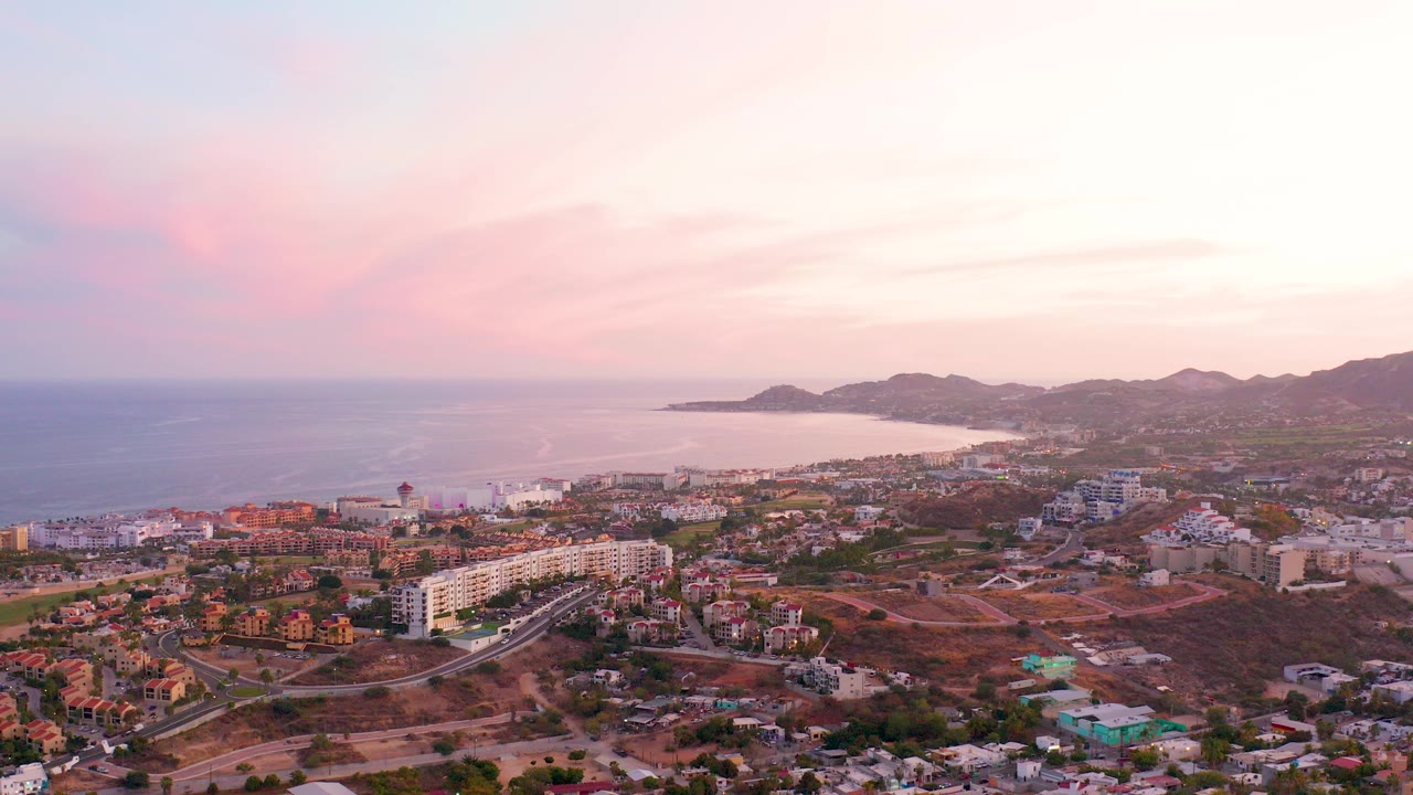 Aerial view over San Jose del Cabo with residential buildings and roads in the foreground, ocean and beach to the left, hills on the horizon, pink sunset clouds, and no visible people.