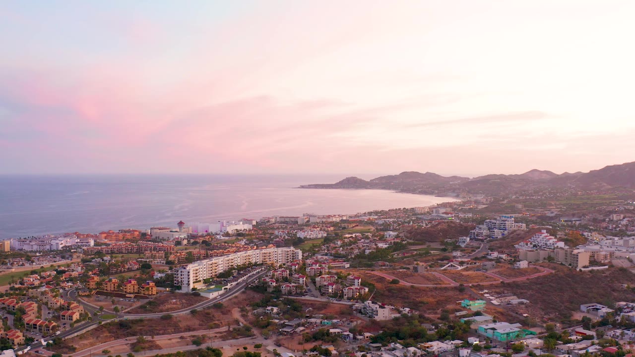 Wide aerial panorama in San Jose del Cabo showing the ocean and beach on the left, hillside residential areas across the lower frame, mountains in the background, and no visible people.