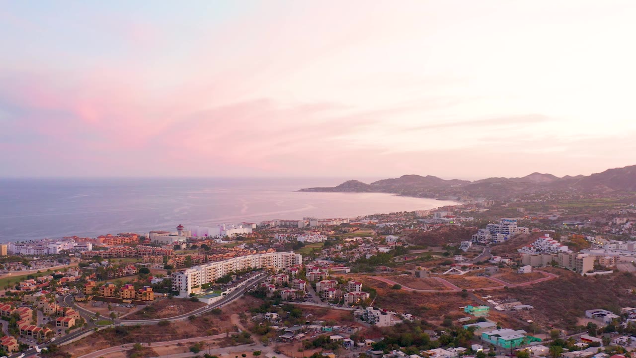 Aerial coastline view with town foreground, ocean left, and hills under pink clouds.