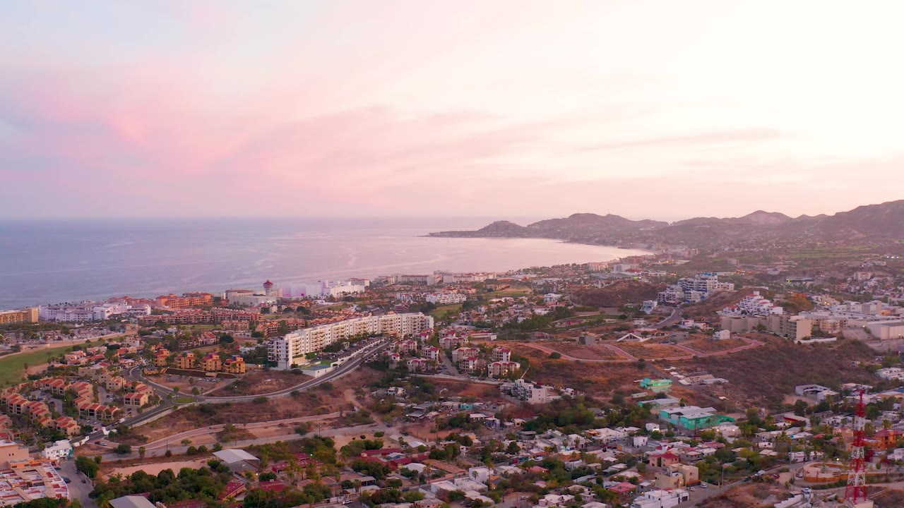 Aerial view across San Jose Del Cabo with white and tan buildings, winding roads, hillside background, ocean to the left, and bright sunset sky, with no visible people.