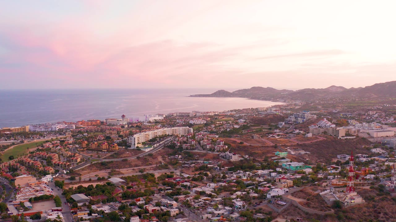 Aerial view over San Jose Del Cabo with low-rise buildings in the foreground, ocean to the left, hills beyond the coast, pink dusk clouds, and no visible people.