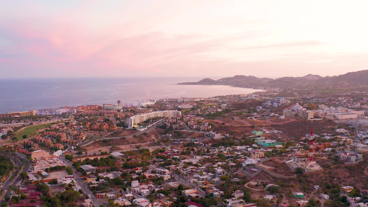 Opening aerial frame over San Jose del Cabo, Mexico, with ocean at left, hillside shoreline in the distance, residential roads below, pink dusk clouds, and no visible people.