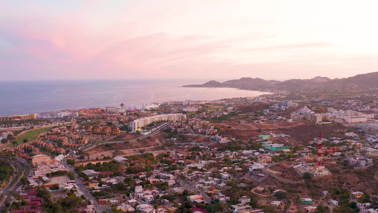 San Jose del Cabo aerial view one second into the clip, showing the ocean and bay beyond low-rise buildings, hillside roads, a large white structure, and no visible people.