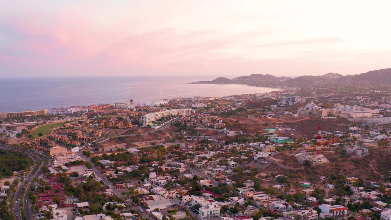 Opening frame of San Jose del Cabo from above, with residential streets in the foreground, ocean on the left, hills in the distance, pink dusk clouds overhead, and no visible people.