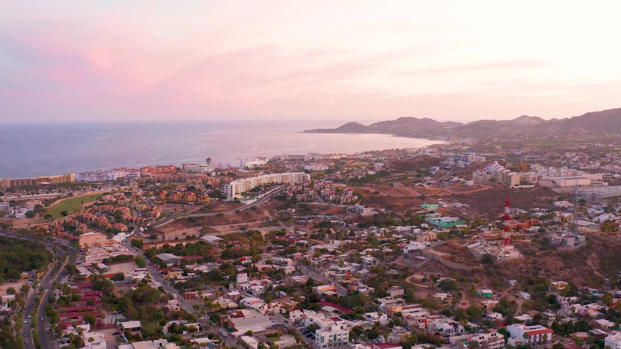San Jose del Cabo remains framed from a high aerial angle, with clustered homes below, a road curving through the lower left, the coastline beyond, and no visible people.