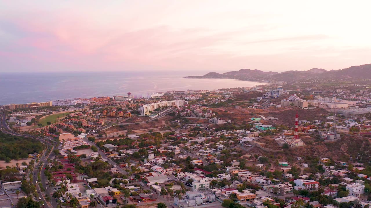 Wide San Jose del Cabo aerial showing residential streets, a church tower near the lower center, coastline, hills, soft pink clouds, and no visible people.