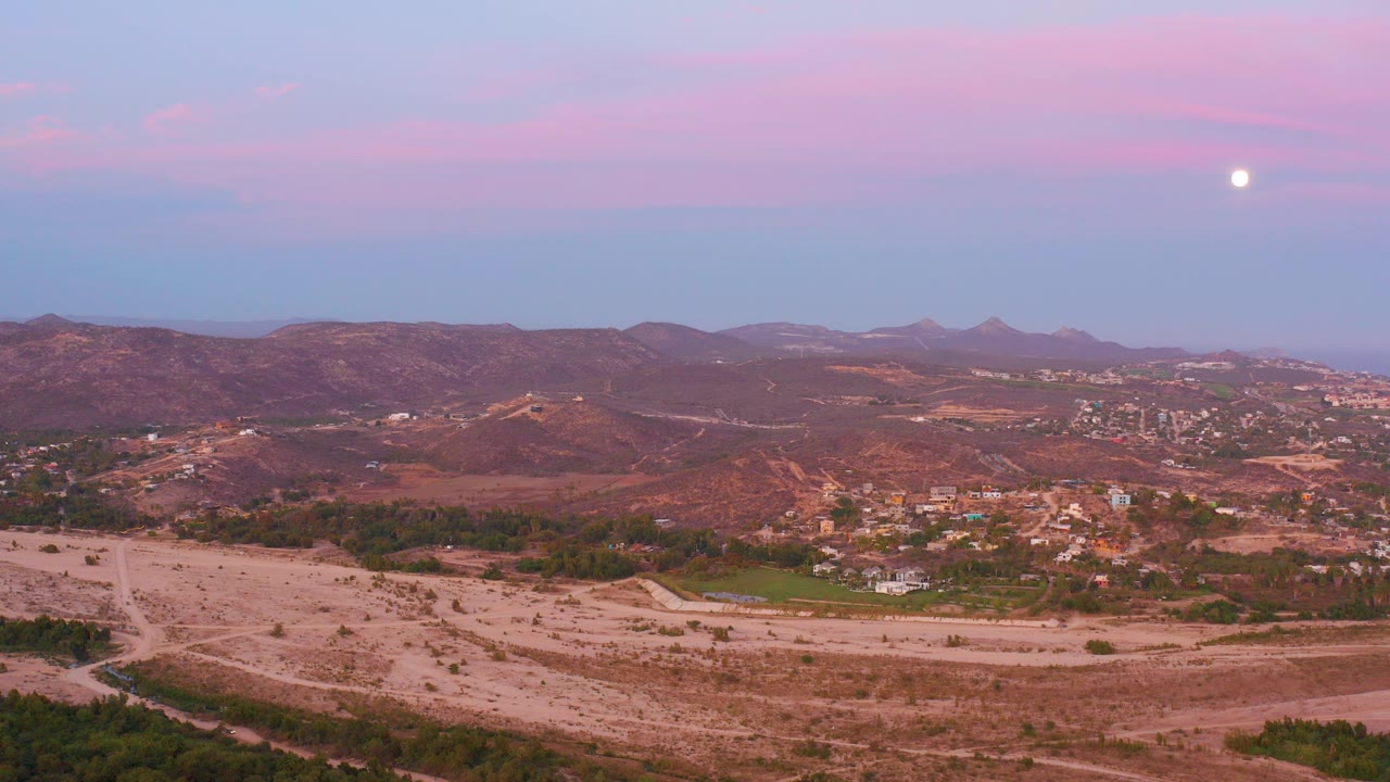 San Jose del Cabo desert valley at twilight with sandy foreground, scattered vegetation, distant hillside buildings, mountains, a bright moon, and no visible people.