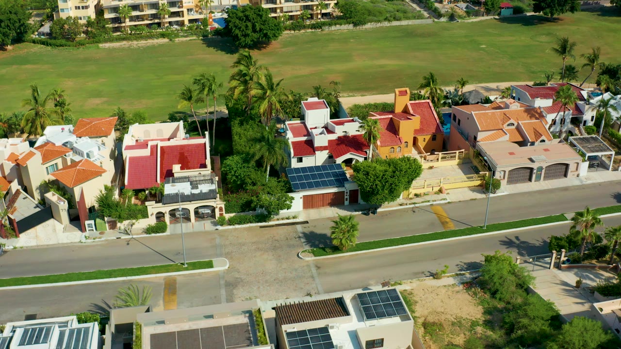 Aerial view in San Jose Del Cabo showing red and orange roofed houses along a road, palm trees, a green field, distant apartment buildings, and no visible people.