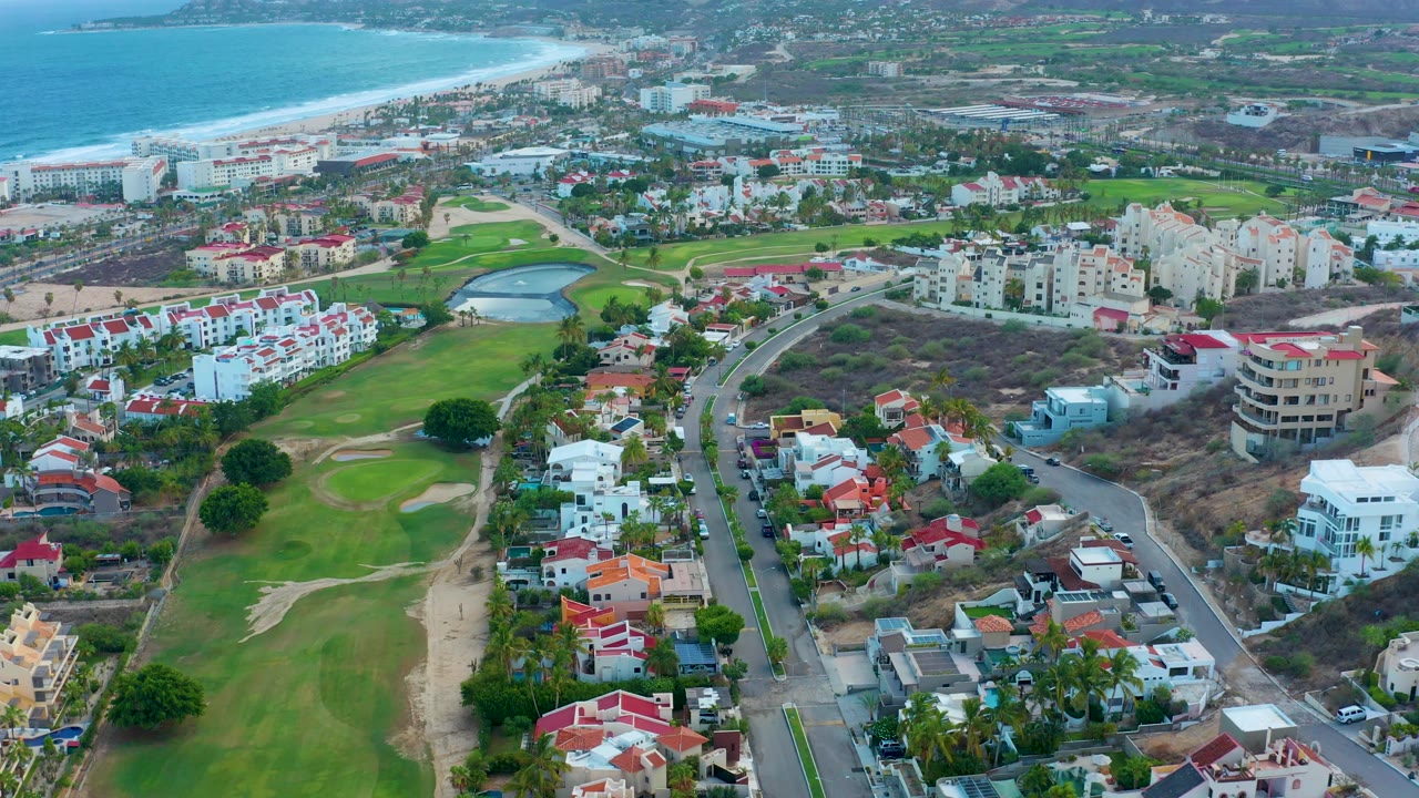 Opening aerial frame over San Jose Del Cabo showing green golf fairways, sand traps, white and red-roofed homes, a curving road, beach, ocean, and no visible people.