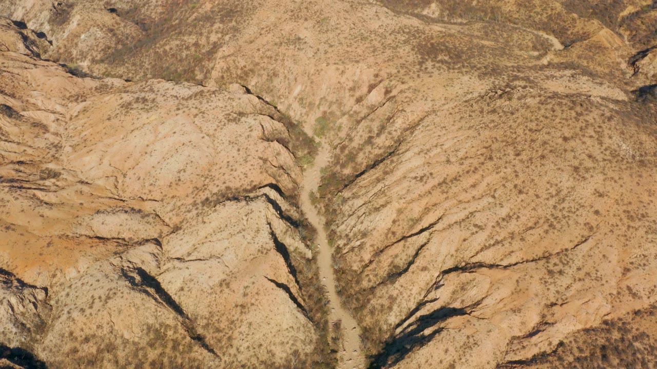 Aerial frame from Cabo Pulmo with the dry channel centered between rounded tan slopes and darker erosion cuts, no visible people.