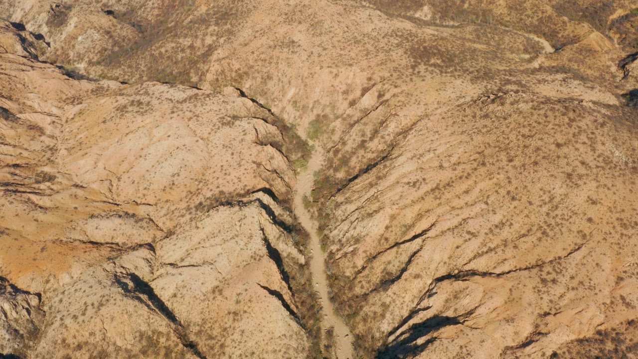Cabo Pulmo aerial view showing a pale wash descending through scrub-speckled beige hills with dark gullies and no visible people.