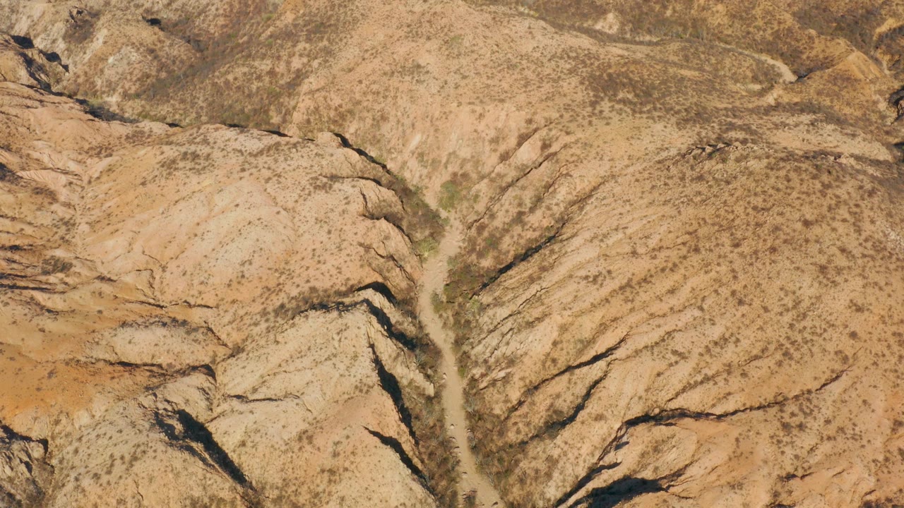 Aerial frame over Cabo Pulmo desert ridges where a narrow dry channel separates tan hills marked by dark rock seams, with no visible people.