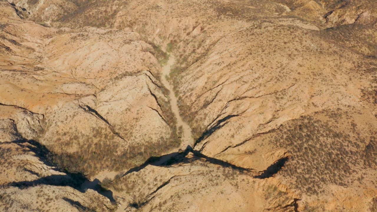 Aerial view over Cabo Pulmo dry canyon terrain with beige ridges, dark erosion cuts, a narrow wash, sparse scrub, and no visible people.