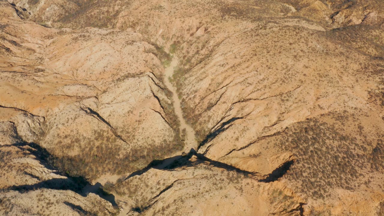 Aerial frame in Cabo Pulmo showing the dry wash more centrally between eroded beige slopes, with scattered desert scrub and no visible people.