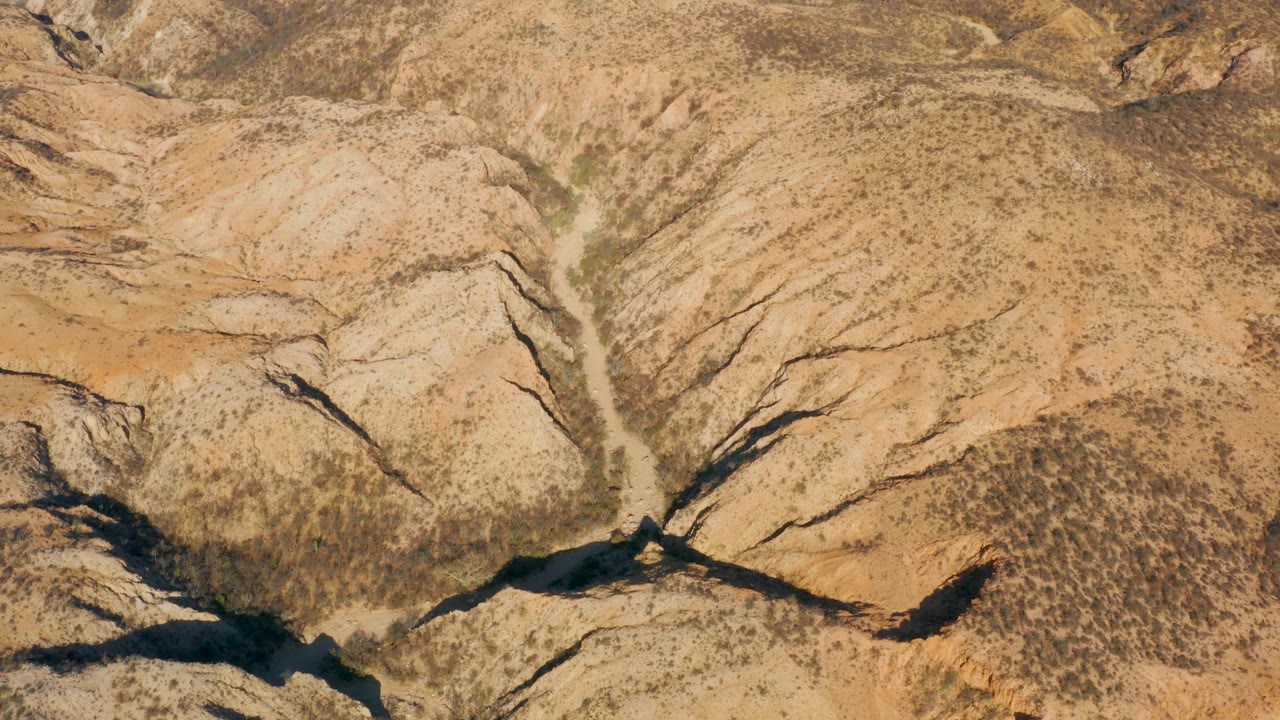 Aerial view of Cabo Pulmo canyon ridges with branching dark gullies, tan rock faces, sparse vegetation, and no visible people.