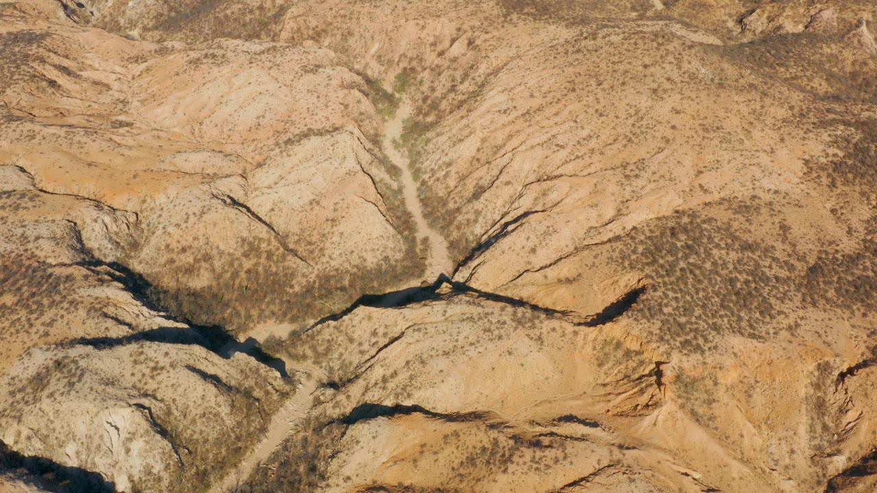 High view over Cabo Pulmo canyon terrain where a pale dirt road or wash runs through tan slopes and dark eroded channels, with no visible people.