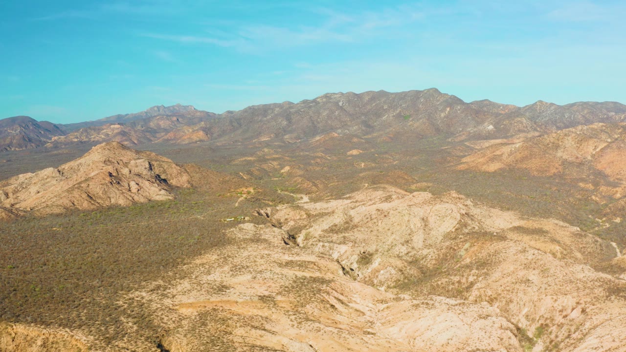One second into the Cabo Pulmo aerial view, tan ridges sit in the lower frame while green-brown scrub stretches toward hazy mountains under a clear blue sky, with no visible people.