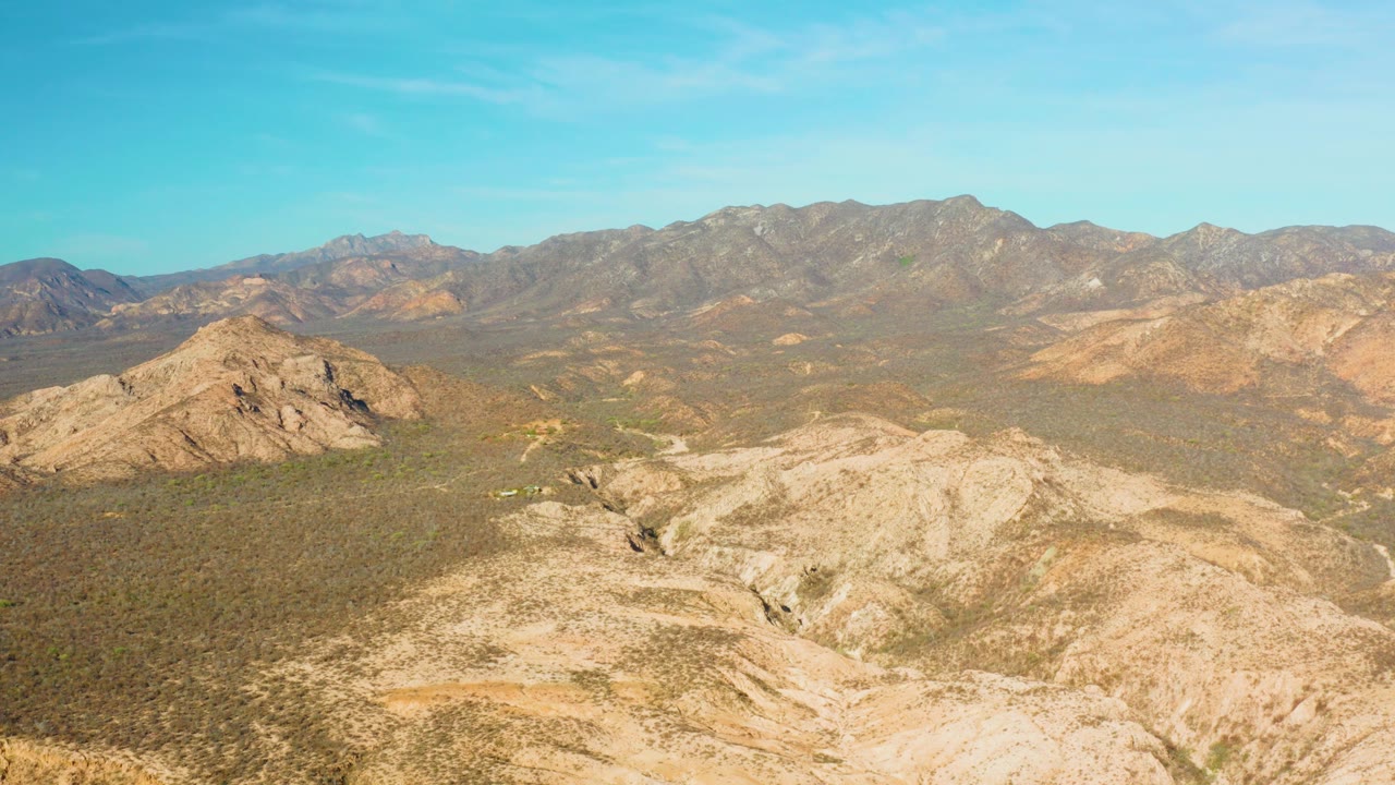 At 0:03, pale foreground rock and sparse vegetation dominate the lower frame, with Cabo Pulmo's inland valley and brown mountain range extending into haze, no visible people.