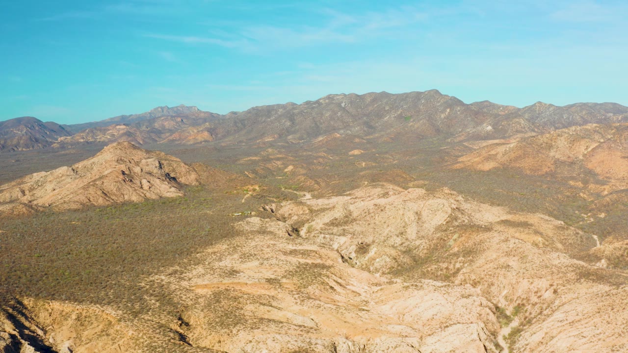 Aerial view in Cabo Pulmo showing pale foreground rock, green-brown desert vegetation, layered mountain slopes in the background, and no visible people.