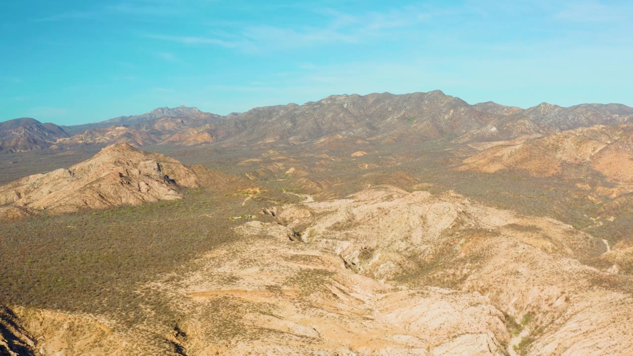 Mid-clip aerial frame over Cabo Pulmo with eroded tan rock in front, scrub-covered flats, dark mountain ridges behind, and no visible people.