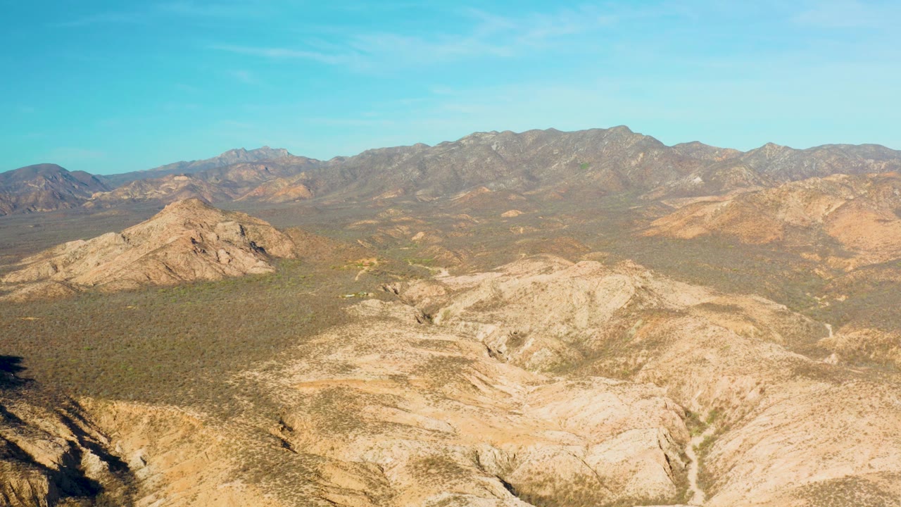 Aerial frame from Cabo Pulmo with tan foreground ridges, a scrubby midground basin, layered mountains on the horizon, and no visible people.