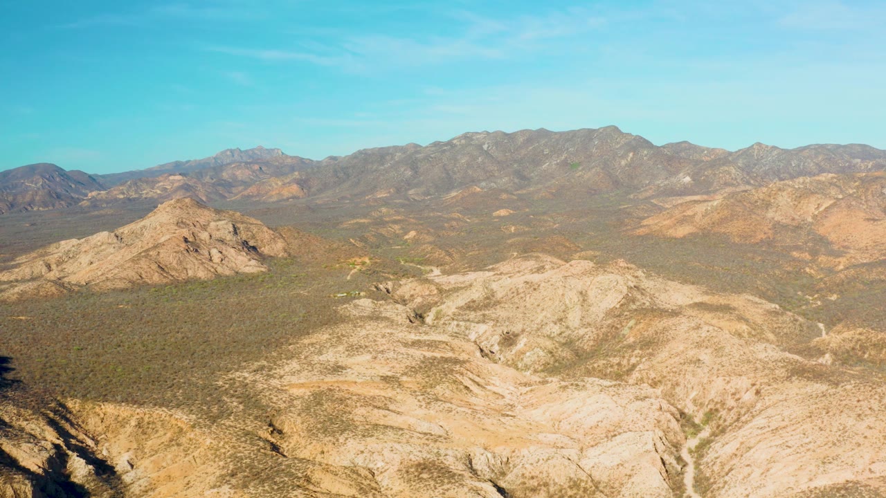 Wide view across Cabo Pulmo desert terrain where dry gullies and ridges fill the foreground, scrub spreads through the valley, and the mountain range sits beyond with no visible people.