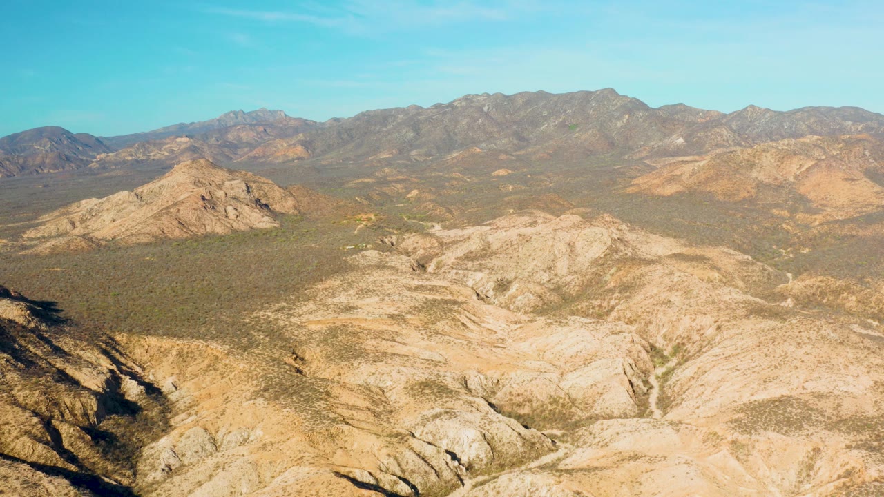Opening frame over Cabo Pulmo, Baja California Sur, with pale rocky washes in the foreground, scrubby valley floor, distant mountains, blue sky, and no visible people.