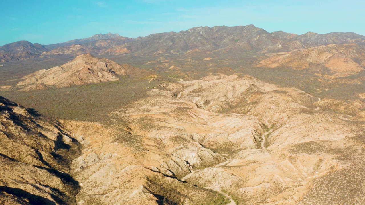 Cabo Pulmo aerial frame with a sunlit rocky basin in front, scrubland across the middle distance, muted mountain ridges on the horizon, and no visible people.