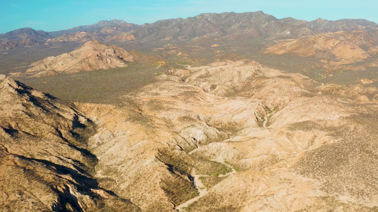 Aerial view in Cabo Pulmo showing pale eroded hills centered below the frame, a scrub valley beyond them, distant mountain ridges, blue sky, and no visible people.