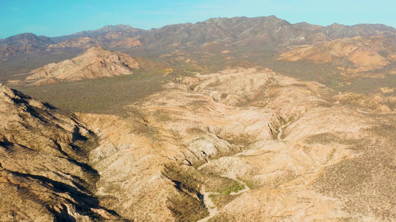 Mid-clip aerial view over Cabo Pulmo with dry ridges crossing the lower frame, muted scrub across the valley floor, distant brown-gray mountains, and no visible people.
