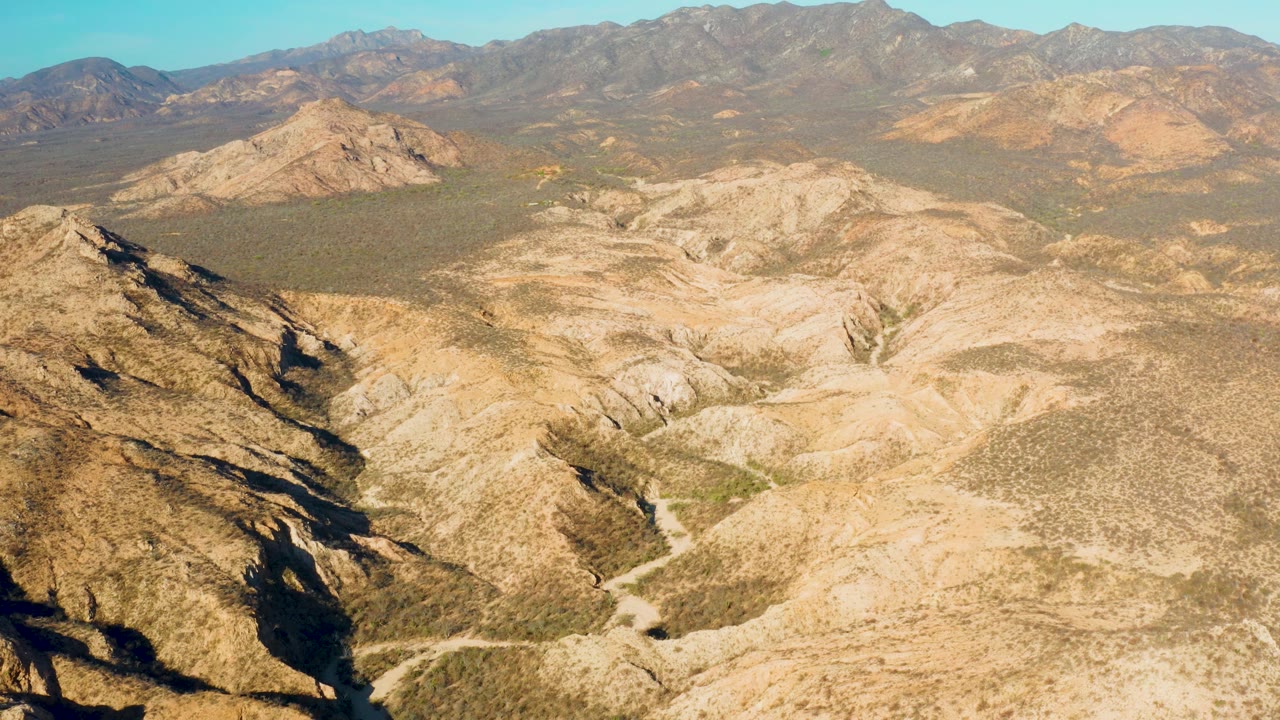 The aerial view continues across Cabo Pulmo rocky hills and dry valley channels, with a shadowed slope at left and broad mountains across the background, with no visible people.