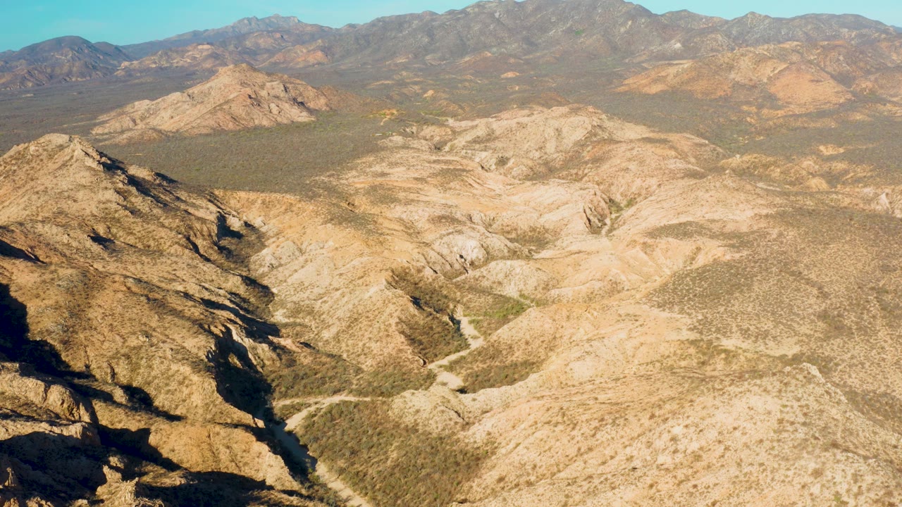 High aerial frame from Cabo Pulmo, Baja California Sur, with beige slopes in the foreground, a winding pale track in the valley, distant ridges, and no visible people.