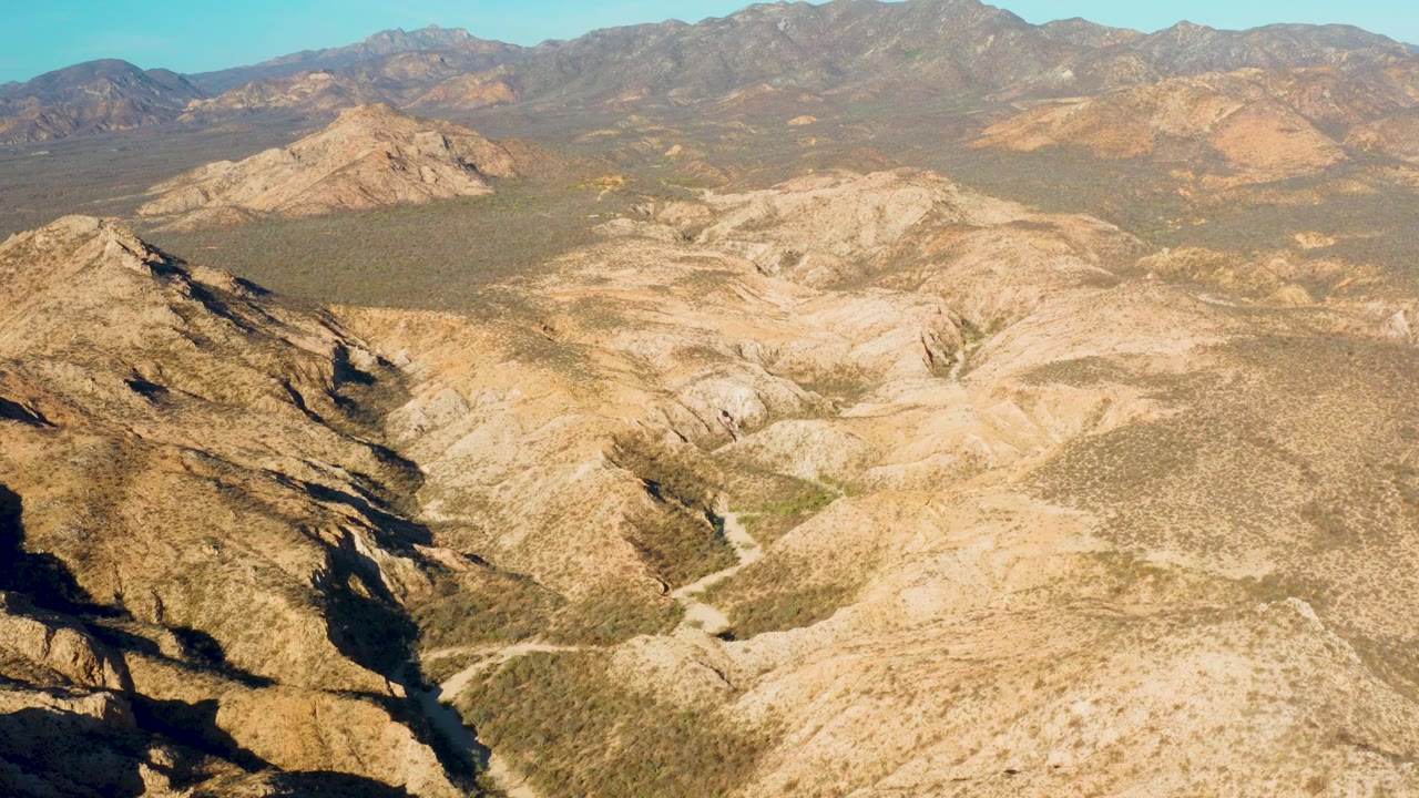 Mid-clip aerial frame over Cabo Pulmo, Baja California Sur, with strong shadows on the left ridge, light beige wash terrain below, distant mountains, and no visible people.