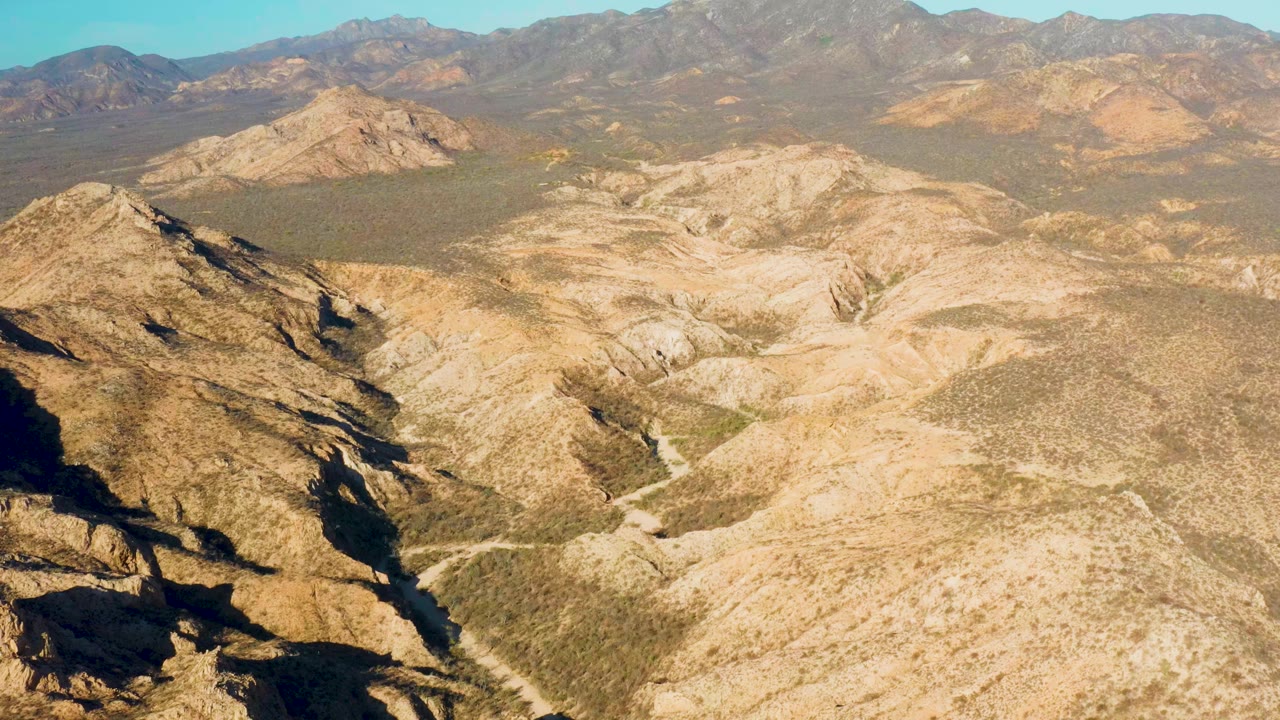 Aerial view over Cabo Pulmo, Baja California Sur, Mexico, showing a dry rocky valley, a pale trail crossing the brush, distant ridges, and no visible people.