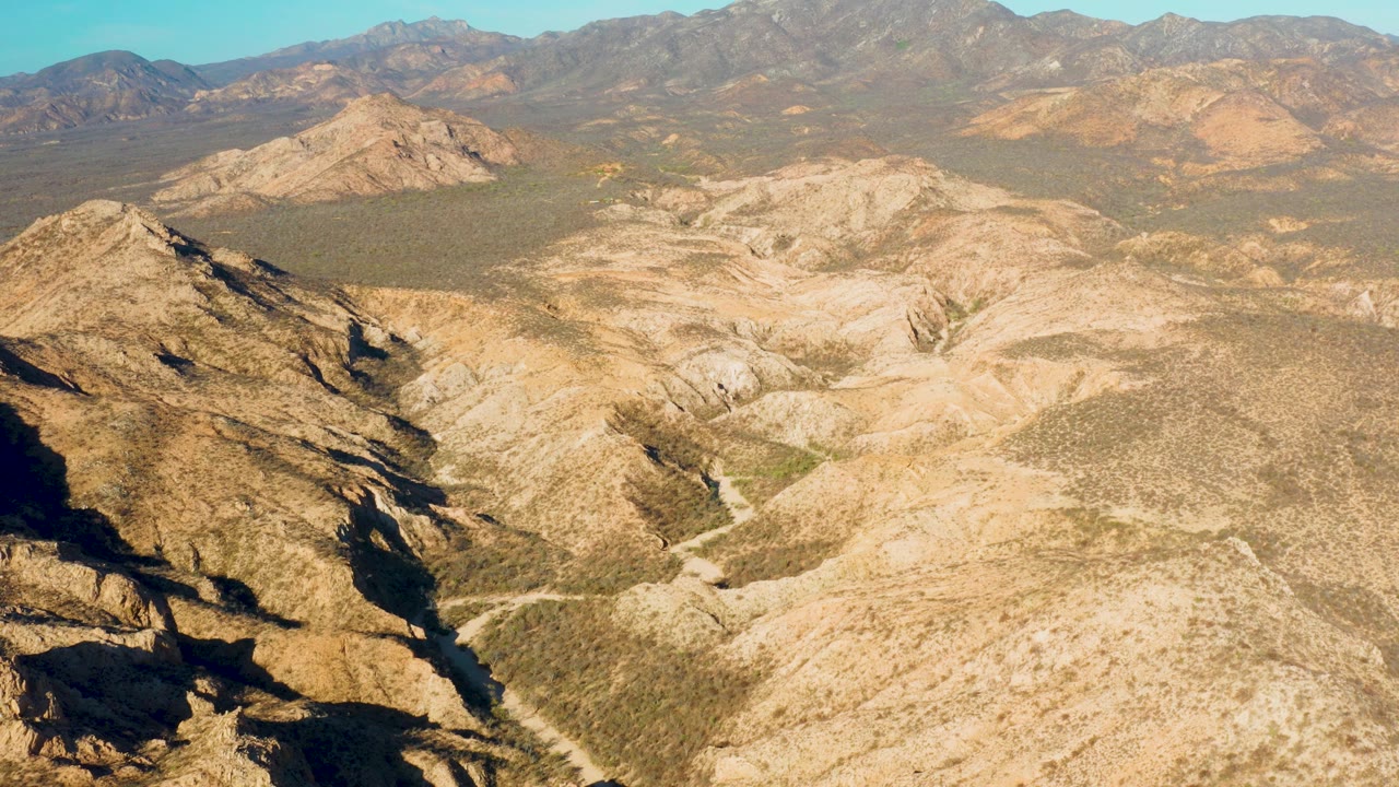 Aerial Cabo Pulmo frame with shadowed rock at the lower left, a dry trail cutting through the valley floor, layered tan hills, distant mountains, and no visible people.