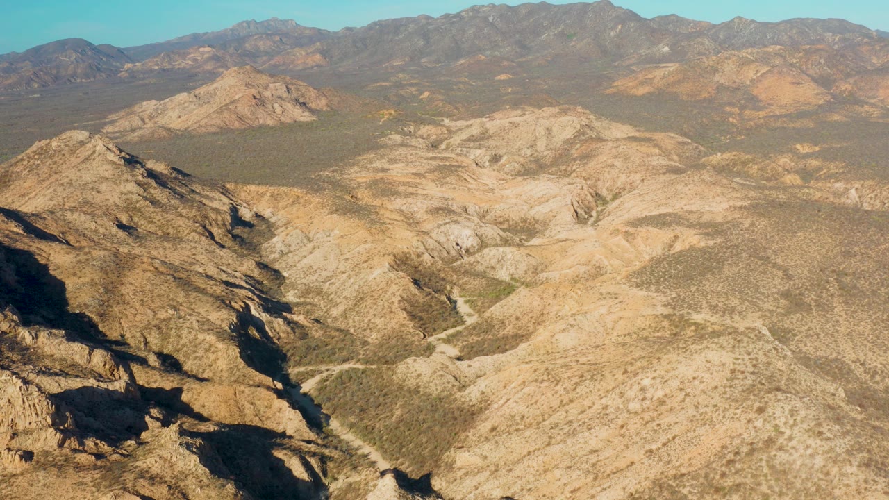 Wide aerial view of Cabo Pulmo rocky desert valleys with tan ridges, sparse scrub, distant mountains, clear blue sky, and no visible people.