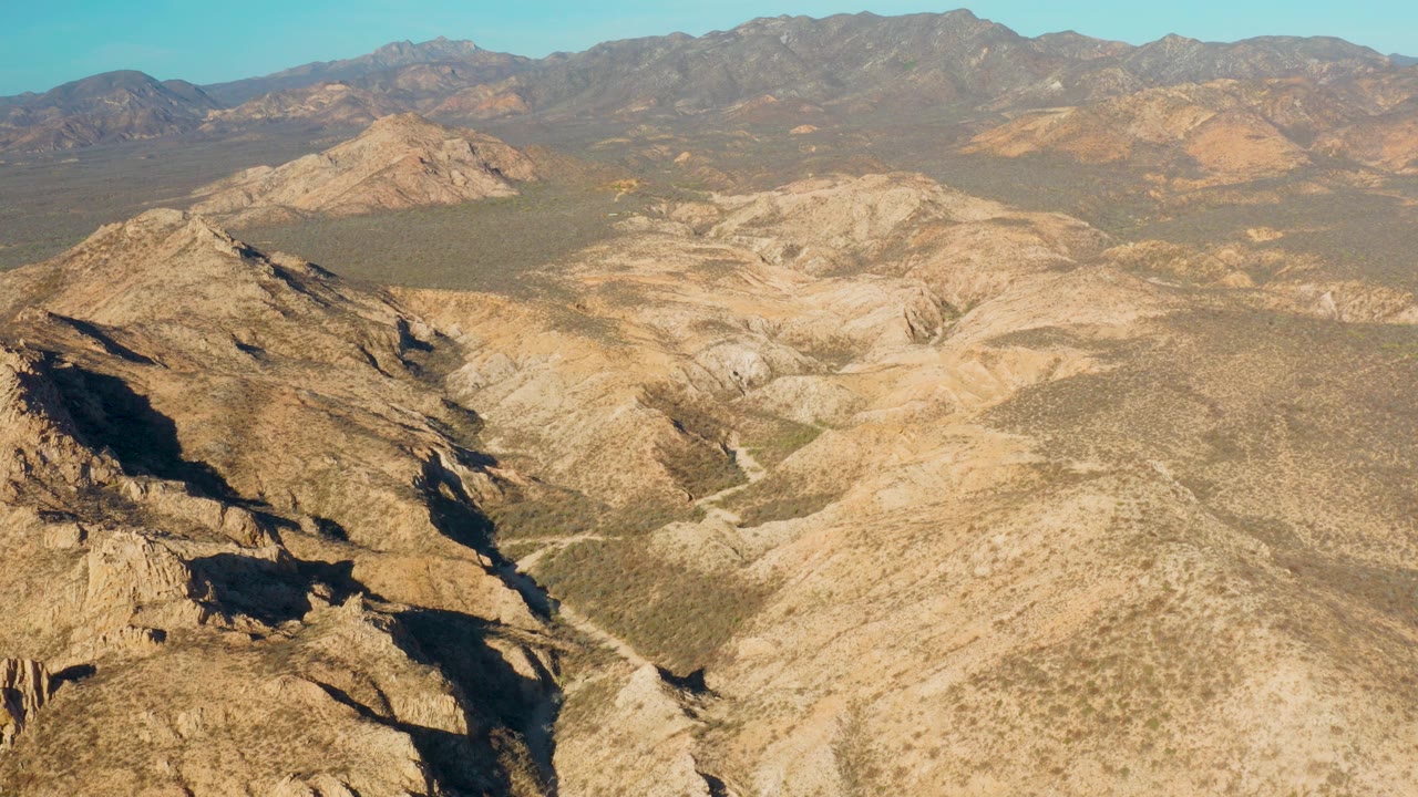 Opening aerial frame over Cabo Pulmo, Baja California Sur, with a shadowed rocky ridge at left, tan desert basin ahead, distant mountains, clear blue sky, and no visible people.