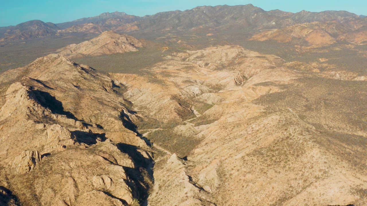 Aerial view over Cabo Pulmo, Baja California Sur, with tan rocky ridges in the foreground, dry valleys in the midground, distant mountains on the horizon, and no visible people.