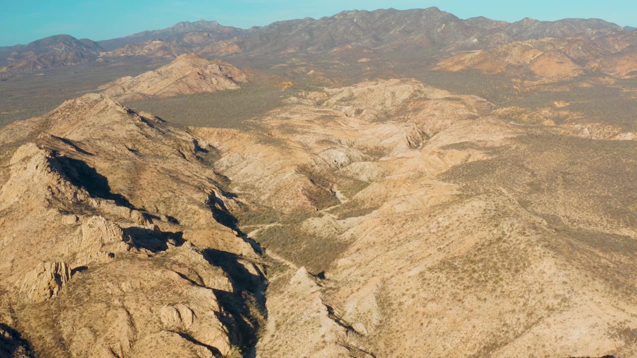 High aerial frame in Cabo Pulmo showing a shadowed rocky ridge at lower left, pale dry washes through the center, layered tan hills, distant mountains, and no visible people.