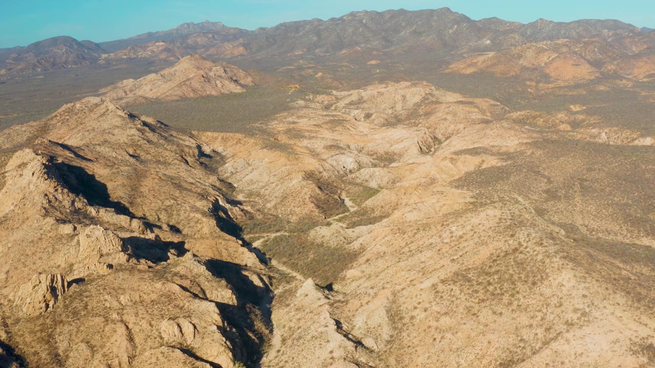 Aerial frame from Cabo Pulmo with a broad dry valley centered between rocky ridges, low desert scrub across tan ground, a mountain range along the horizon, and no visible people.