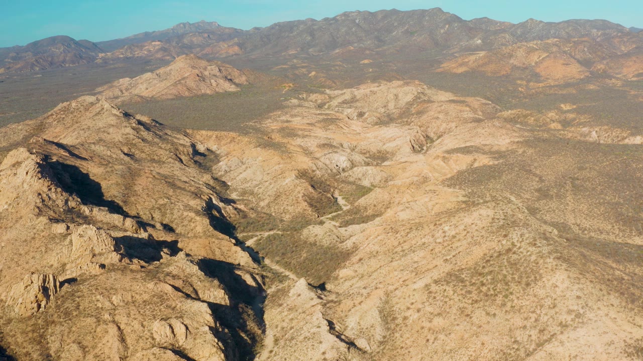 Sunlit desert hills with dark ridge shadows and distant mountains under clear blue sky.