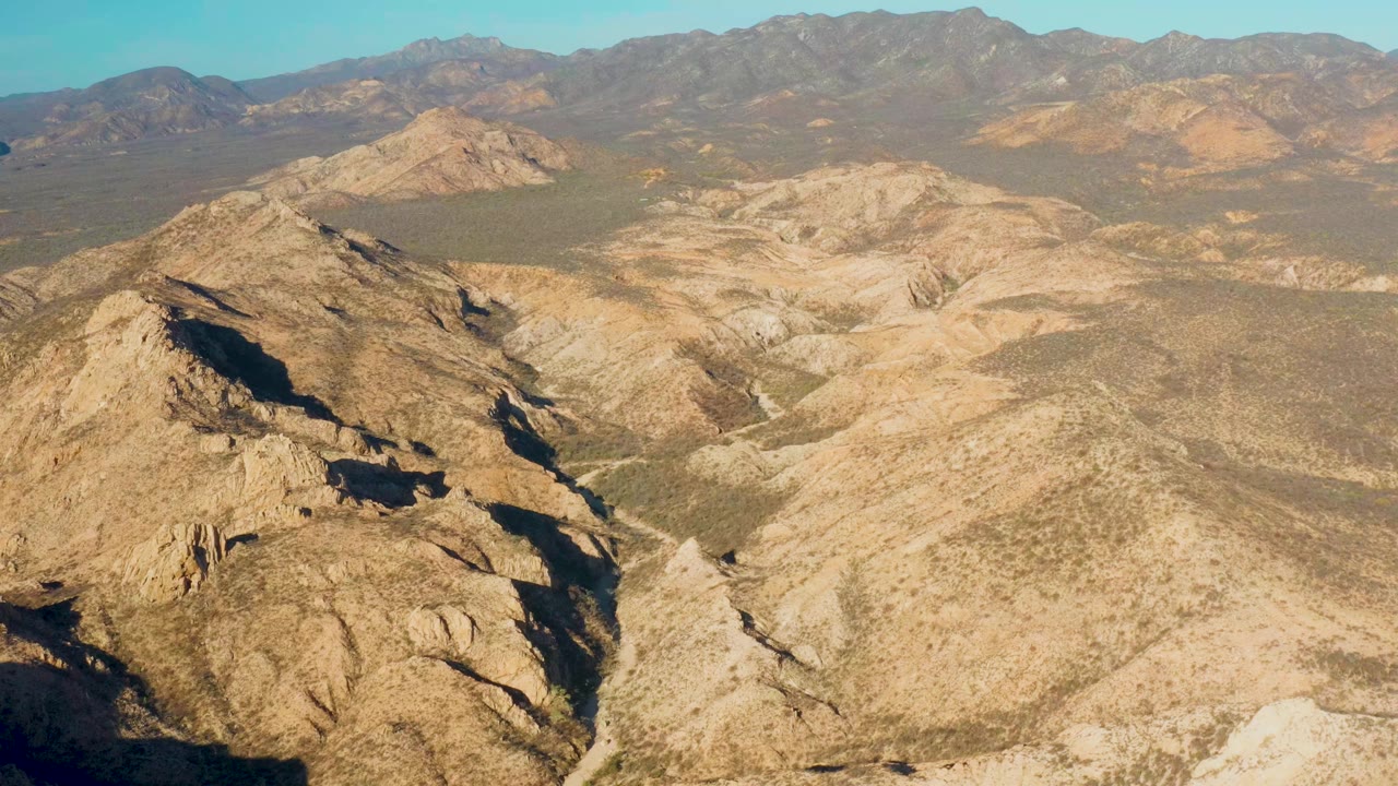 High aerial view over Cabo Pulmo, Baja California Sur, with tan rocky ridges in the foreground, pale arid hills in the center, distant mountains on the horizon, and no visible people.