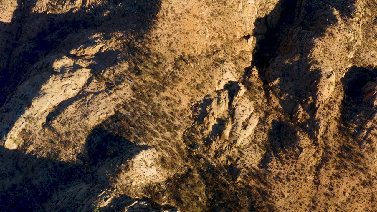 Overhead view of rocky arid terrain in Cabo Pulmo, Baja California Sur, with tan slopes, sparse scrub, deep shadows at the left edge, and no visible people.