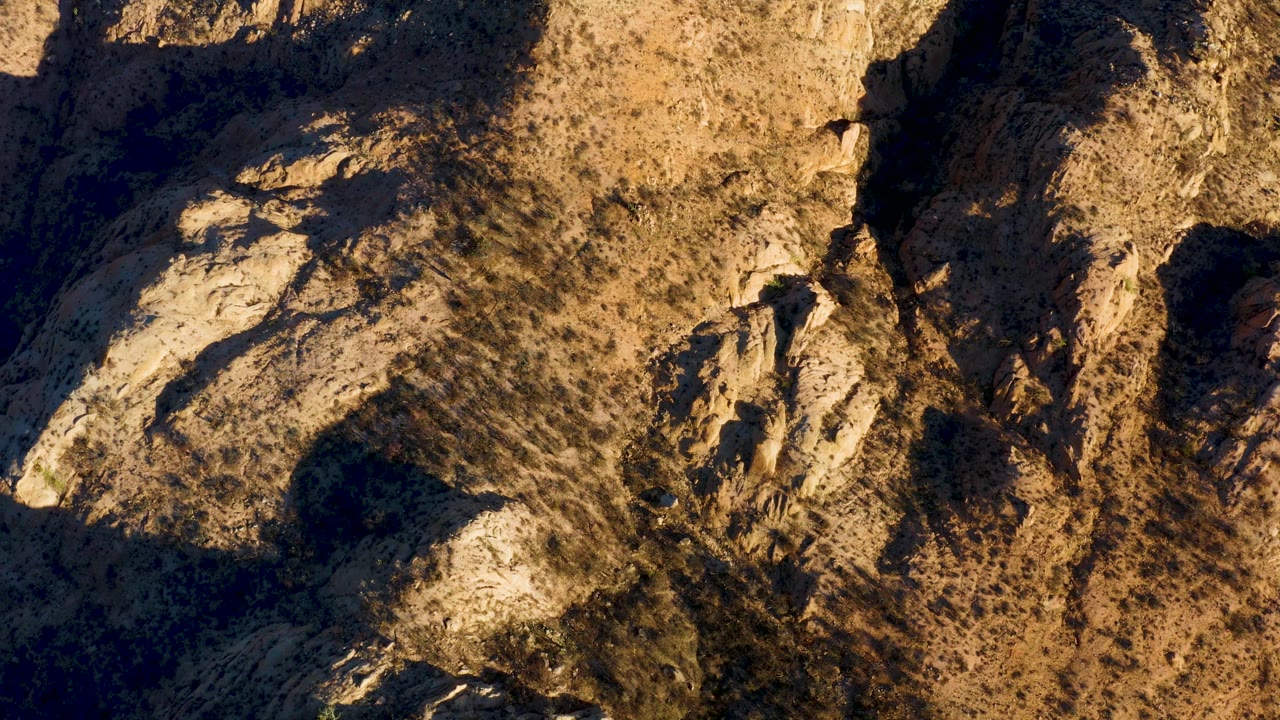 Aerial frame of beige rock and dry scrub in Cabo Pulmo, Baja California Sur, with a broad sunlit ridge and dark gullies cutting through the terrain, no visible people.