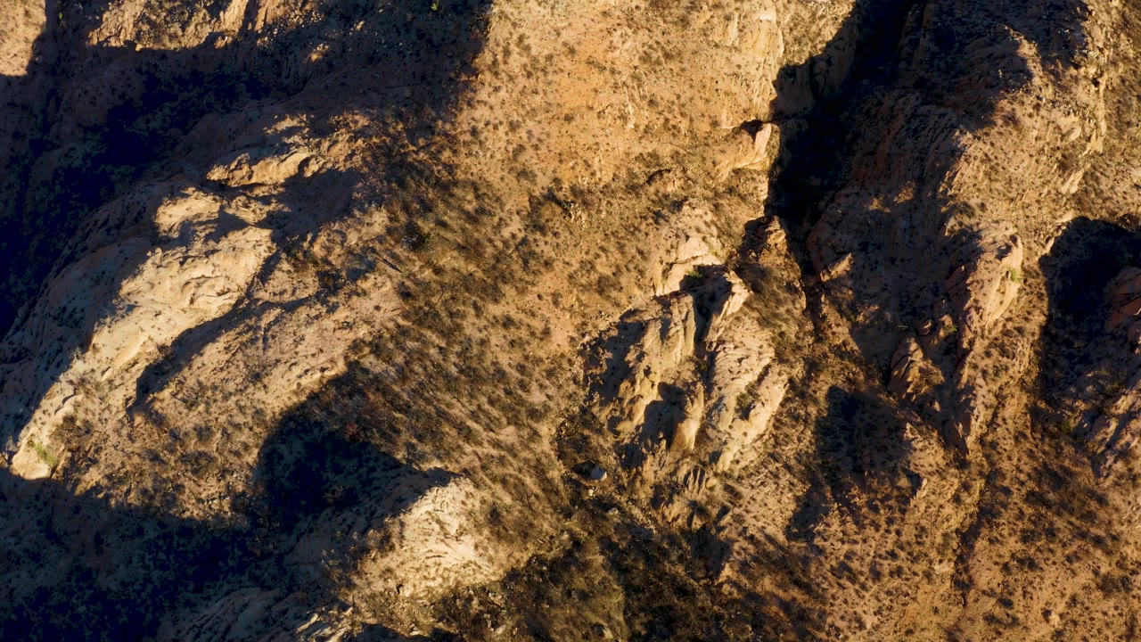 Overhead Cabo Pulmo terrain with tan rocky ground, scattered brush, a bright rock face near the lower left, and heavy shadow along the right side, no visible people.