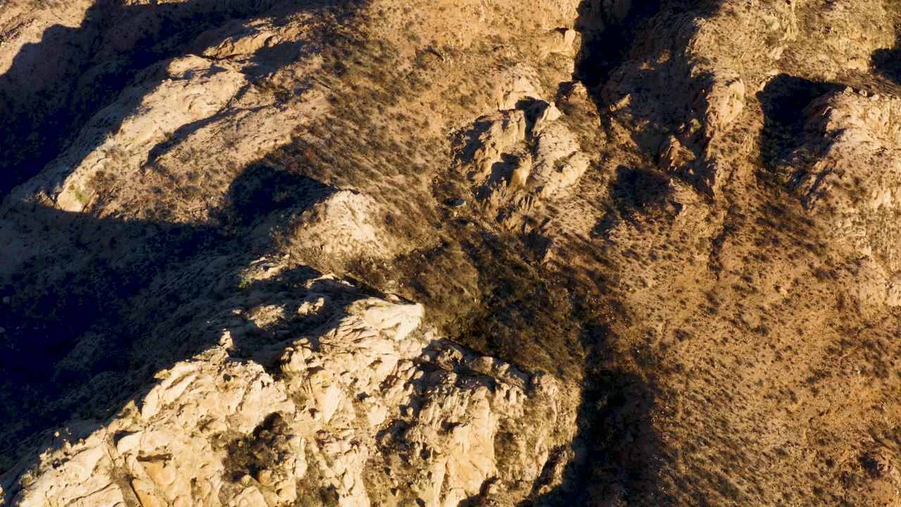 Cabo Pulmo hillside detail showing tan ridges, pale rock shelves, scattered dry scrub, shadowed cuts in the terrain, and no visible people.
